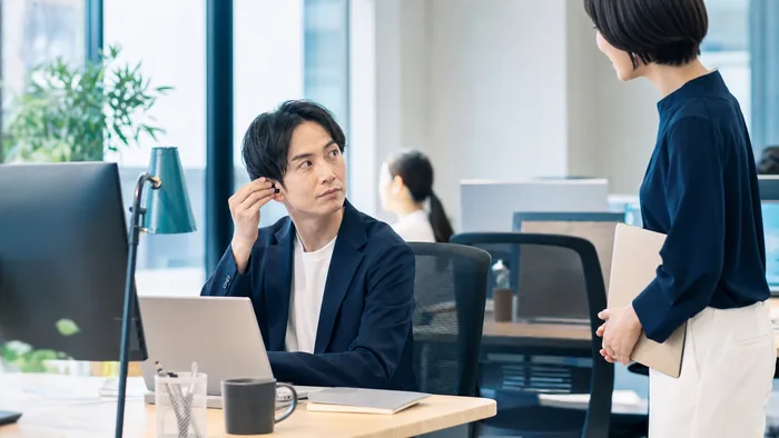 オフィスで同僚に話しかけられ片耳のイヤホンを外す日本の会社員