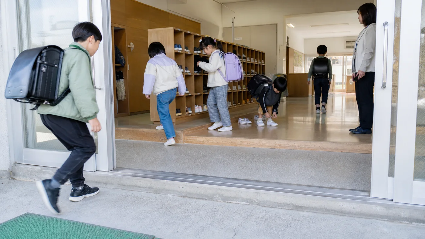 日本の小学校の明るい昇降口で、児童が段差の手前で外靴を脱ぎ、下足箱から上履きを取り出し、教師が見守る様子